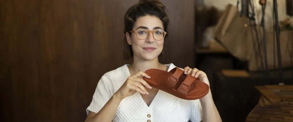 Isabela Chusid premiada por lançar a primeira sandália de plástico vegana. Ela é branca, usa óculos marrom claro, veste uma blusa branca. A sandália em sua mão é da cor marrom.