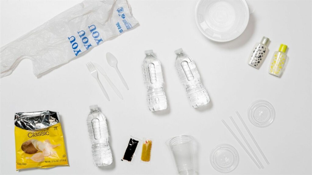 Na imagem aparece materiais que usam o plástico como base. Entre os objetos estão: garrafas plásticas, embalagens de batata frita, sacolas, recipientes de cosméticos, vasilhas, copos e talheres
