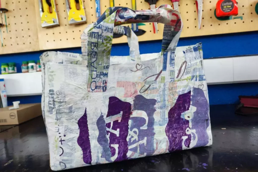 Na imagem aparece uma ecobag feita de sacolas plásticas. Ela é branca com cores roxas 